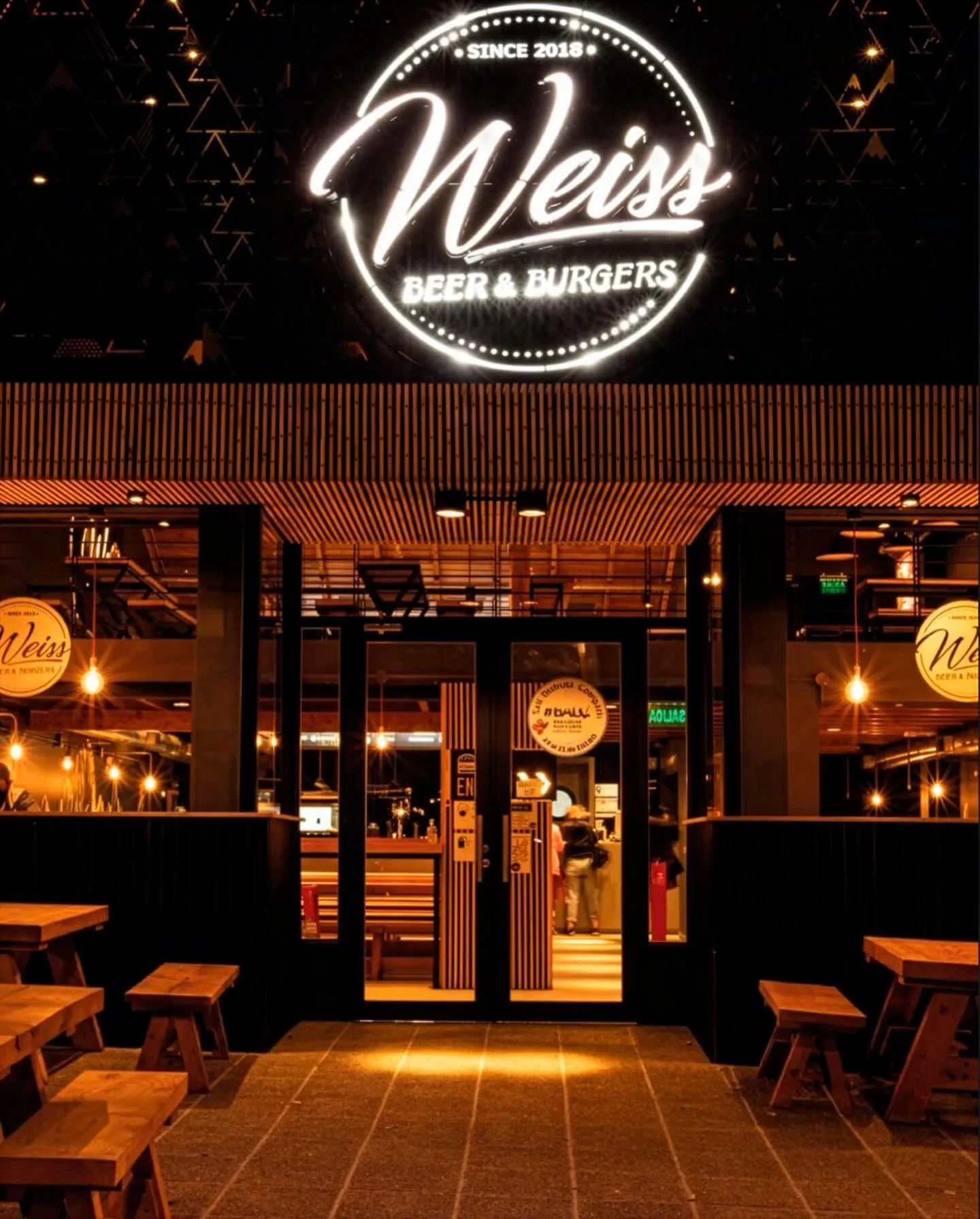 Fachada de noche de un local Weiss con cartel de neón Weiss Beer & Burgers y mesas de picnic