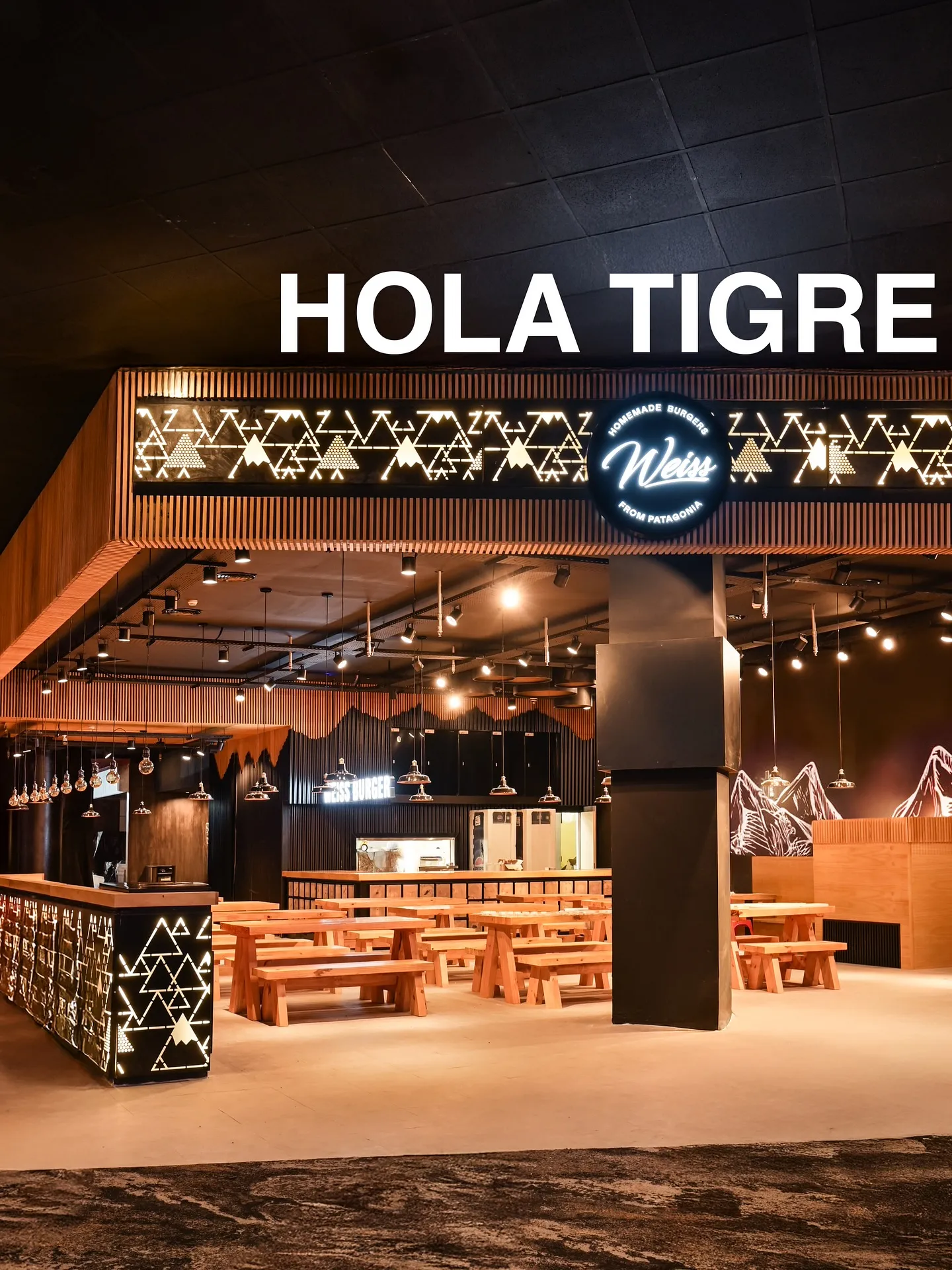 Salón interior de Weiss con mesas de madera, luces cálidas y pared con silueta de montañas